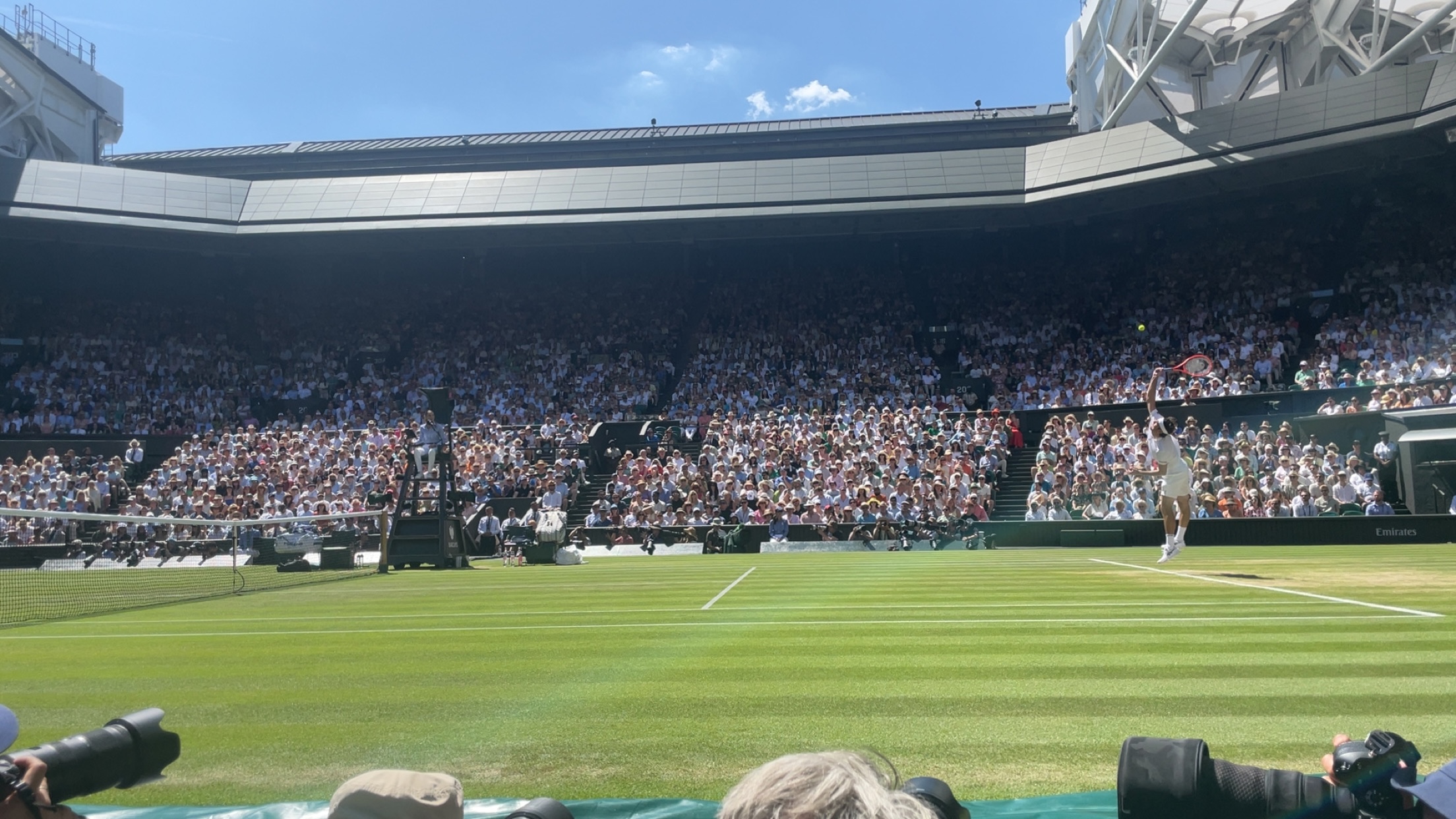 Tennis player serving on a grass court at Wimbledon in front of a packed crowd.